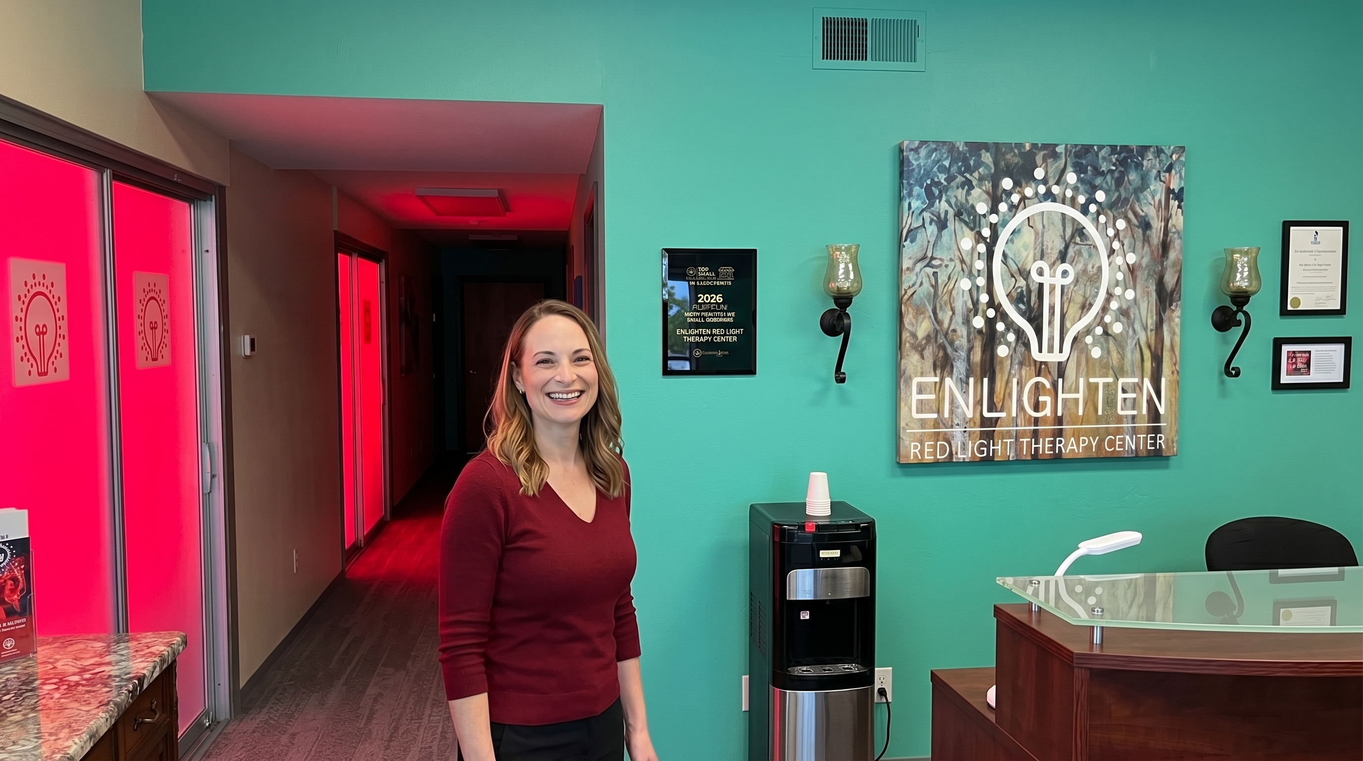 Jennifer Sharp, owner of Enlighten Red Light Therapy Center in Sacramento, smiling in her studio reception area, with the studio's bulb logo and Enlighten Red Light Therapy Center signage on the teal back wall, an award plaque next to her, and warm red light glowing from the treatment hallway behind.